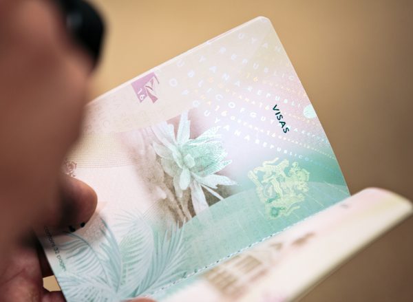 Hands hold a Guyanese passport open to a Visa page with green and yellow artwork and intricate inscriptions