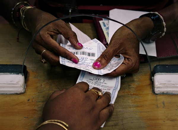 Hands with brightly coloured nails extend through glass opening to submit lottery ticket to attendant